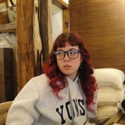 the image of a woman with red hair and glasses sitting at a cafe in a grey hoodie with the word “yonsei” on it