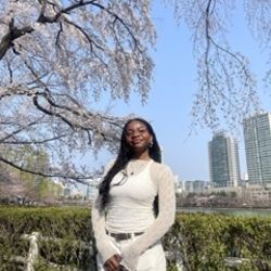 Promise standing in front of a cherry blossom tree and greenery, and set in the background is a lake and 2 tall buildings.