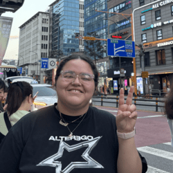Blog writter Maya, posing with a peace sign in front of a 12-story Daiso. Wearing a black t-shirt with a metallic/white start design and a big smile on her face!