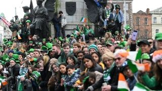 A crowd of people waiting on the parade, dressed in green Irish emblems and Irish scarves. There is a big crowd of people. They are on O'Connell Street looking at the Camera.