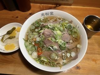 picture of a bowl pho with a plate of peppers to the left and a cup of tea to the right of it
