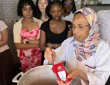 A woman provides instructions to students while adding ingredients to a pot