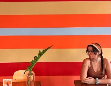 A girl in a headband and glasses sits alone a table in a cafe against an orange striped background.