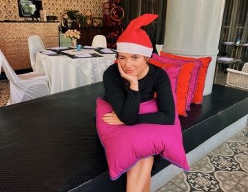 A girl in a black shirt wears a Santa hat and holds a pink pillow in a fancy restaurant