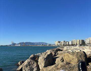 A person's legs are visible as they lounge on a beach jetty, looking out at a view of the water and the city in the distance.
