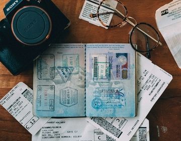 Photo of a passport with visa stamps on a desk scattered with a camera, glasses, and old flight boarding passes