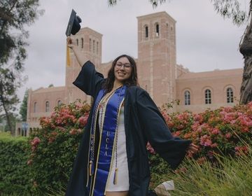 graduation photo, woman in front of university building raising graduation cap in air 