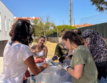 A group of volunteers wearing gloves sort colorful plastic bottle caps into large bins at an outdoor table on a sunny day.