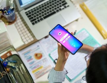 A teenage student interacts with an AI chatbot on a smartphone while studying at a desk with a laptop, notes and stationery. 