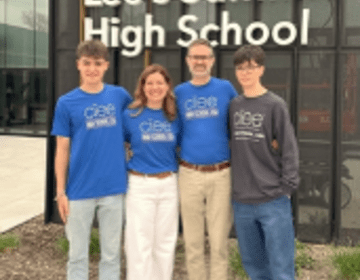 two male students and their host parents wearing with blue shirts with the ciee logo and standing in front of a "lee's summit high school" sign 