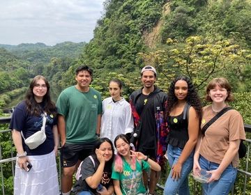 A group of 8 students (mixed gender) smiling and standing in front of a forest/mountain background