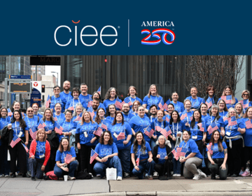 A group of ~50 people in blue "CIEE" standing in a metropolitan area holding american flags and smiling