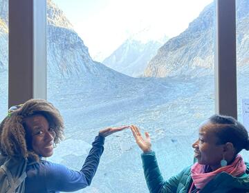 Jemise Ray De La Cruz and her sister smile and gesture toward the alpine landscape visible through a large window overlooking the Mont Blanc mountain range.