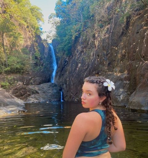 Girl with flower in her hair, wades in the water in front of a waterfall.