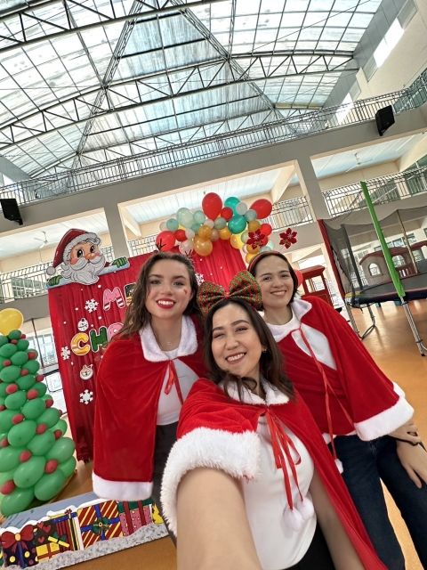 3 women dressed up for Christmas pose for a selfie in front of a Christmas backdrop