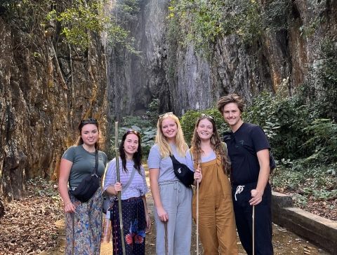 5 people hold walking sticks and smile in front of an opening of a cave with tall trees in the background