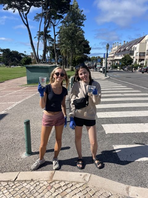 Two students stand smiling by the road holding clear plastic cups containing cigarette butts in blue gloved hands.