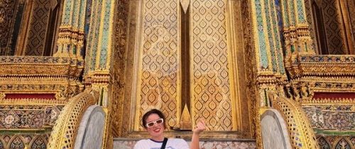 A young woman in a white shirt and colorful pants poses with a mini-heart in front of a gold and blue temple at the Grand Palace in Bangkok, Thailand