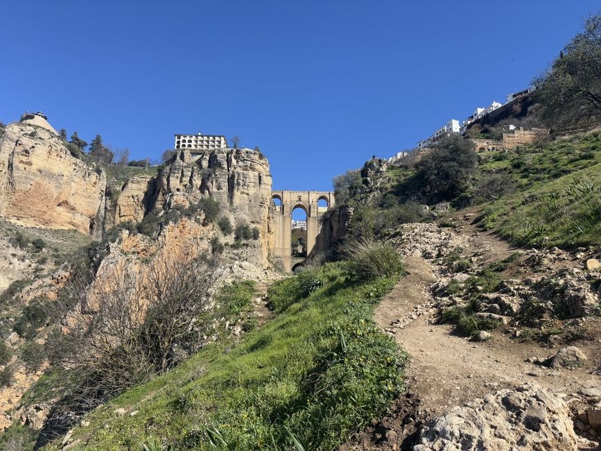 A view of El Puente Nuevo in Ronda, Spain, from below. A rocky trail rises toward the imposing bridge on the right side of the picture. The bridge looms between two cliffs.