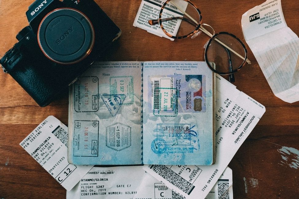 Photo of a passport with visa stamps on a desk scattered with a camera, glasses, and old flight boarding passes