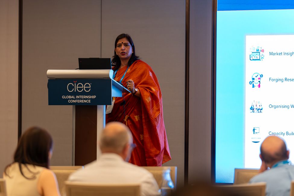 a woman wearing a sari (indian gown) standing behind a podium addressing a seated crowd