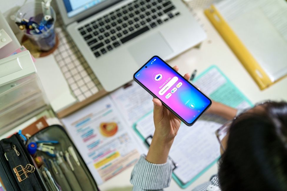 A teenage student interacts with an AI chatbot on a smartphone while studying at a desk with a laptop, notes and stationery. 