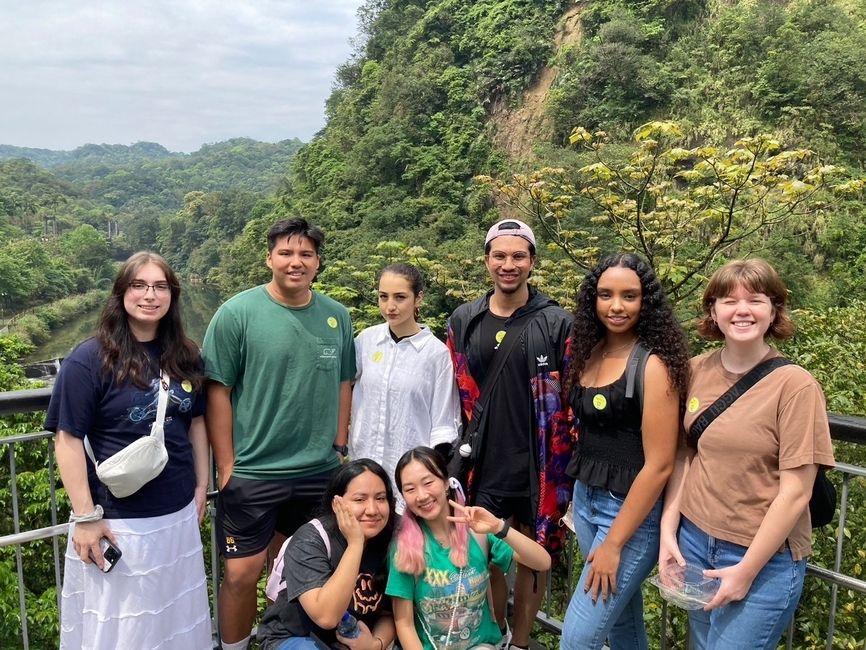 A group of 8 students (mixed gender) smiling and standing in front of a forest/mountain background