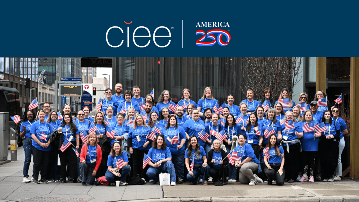 A group of ~50 people in blue "CIEE" standing in a metropolitan area holding american flags and smiling