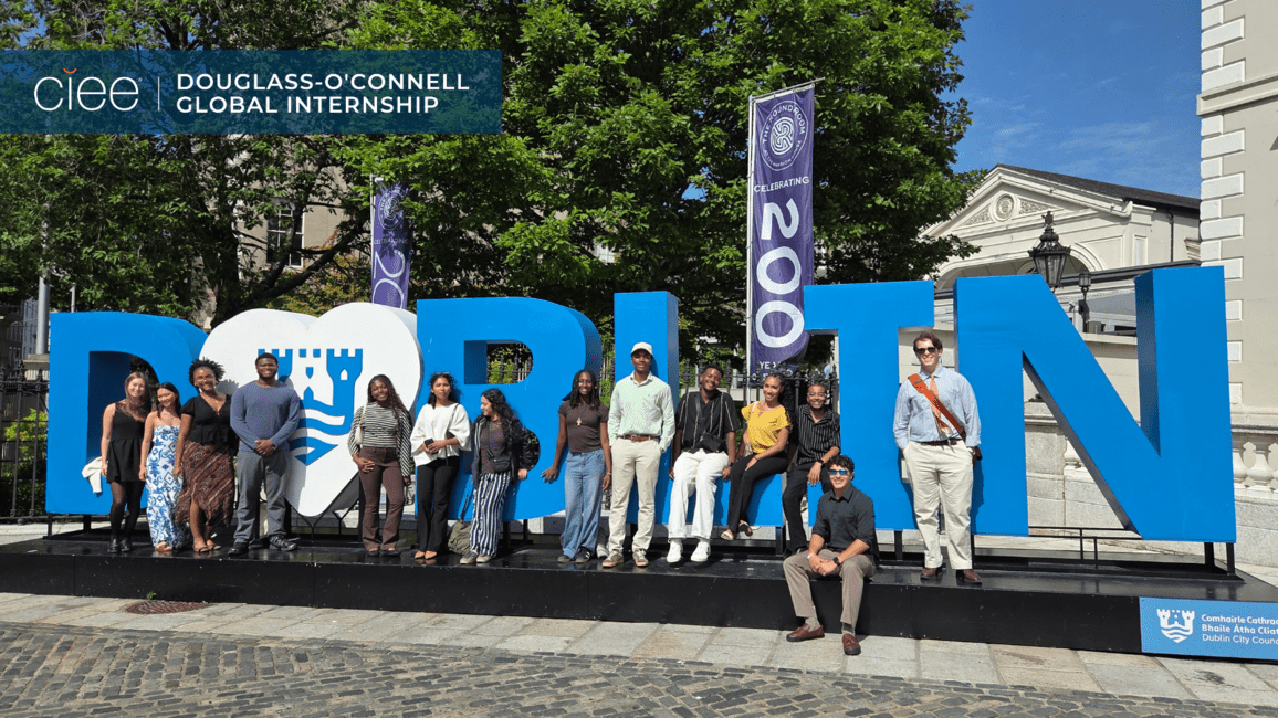 The interns pose around and in front of Dublin’s large blue “DUBLIN” city sign in a public square on a sunny day. Trees, banners, and historic buildings frame the scene. Some students stand while others sit on the letters, creating a relaxed and energetic group portrait. The image highlights the cohort’s arrival in Ireland and captures the vibrant atmosphere of the city where they will live and work during the program.