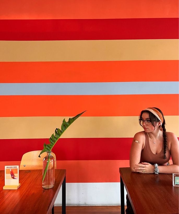 A girl in a headband and glasses sits alone a table in a cafe against an orange striped background.