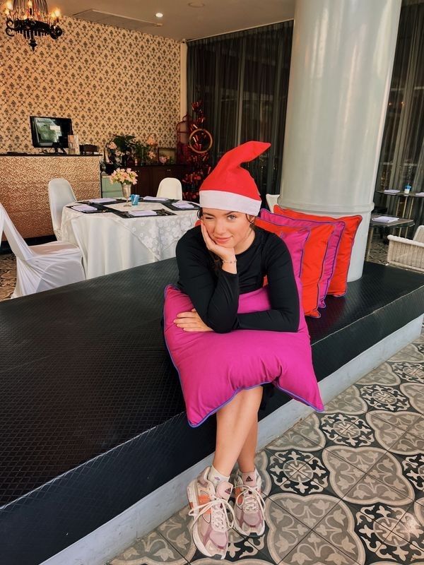 A girl in a black shirt wears a Santa hat and holds a pink pillow in a fancy restaurant