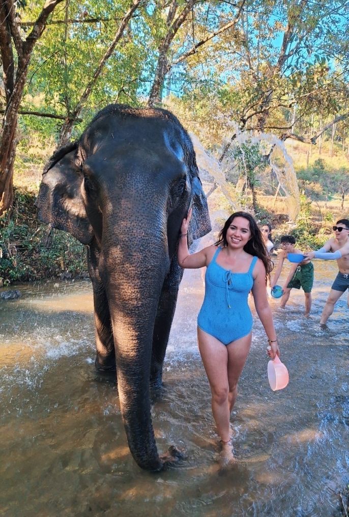 a young woman in a blue swimsuit smiles with an elephant in a creek as people throw water onto the elephant in the background