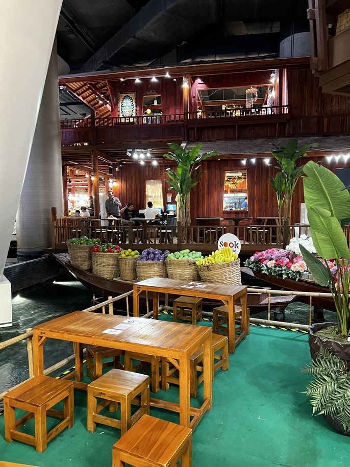 a table with chairs in front of a boat filled with colorful fake fruits in the icon siam food court in bangkok