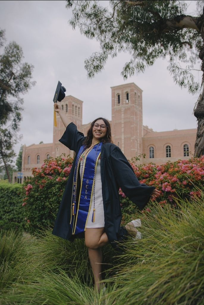 graduation photo, woman in front of university building raising graduation cap in air 