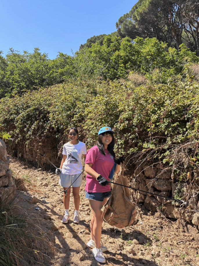 Two smiling high school volunteers carry litter pickers and a collection bag during a cleanup along a rocky trail surrounded by trees and shrubs.