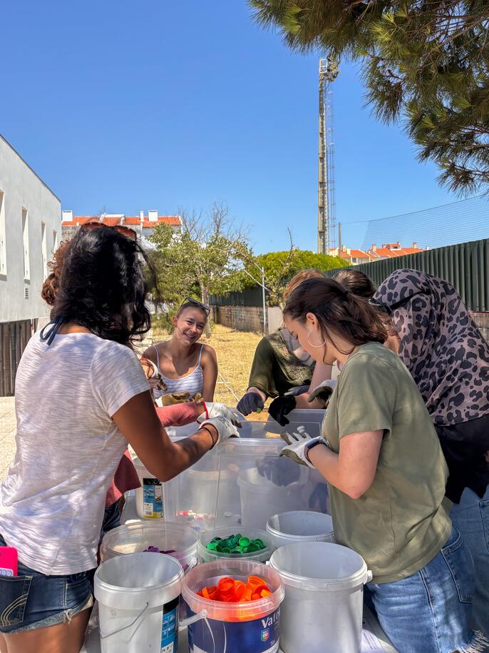 A group of volunteers wearing gloves sort colorful plastic bottle caps into large bins at an outdoor table on a sunny day.