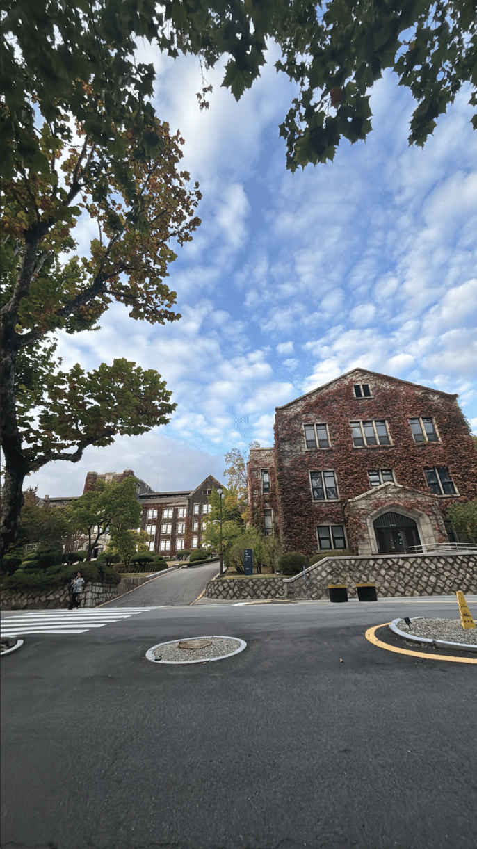 A photo of one of the academic buildings on Yonsei University's campus. The building is covered in vines and leaves turning into fall colors, with cloudy bright blue sky in the background.