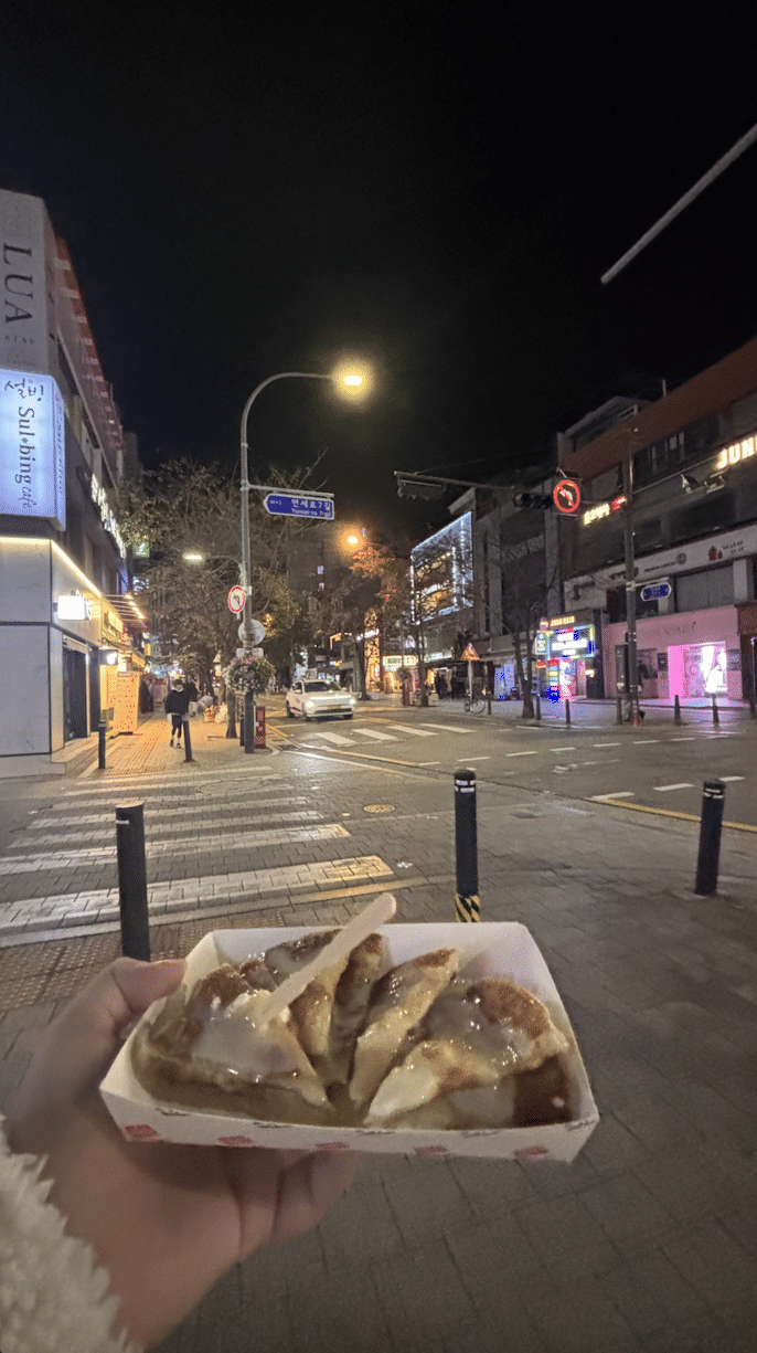 A 0.5 photo of a hot batch of hotteok at night, with the lights of Sinchon painting the background. 