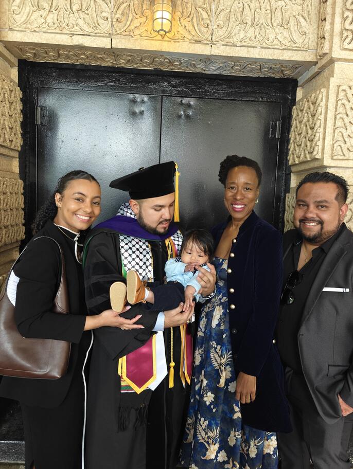 Jemise Ray De La Cruz smiles with her family after a graduation ceremony. A graduate in academic regalia holds a baby while family members gather together in front of an ornate building entrance.