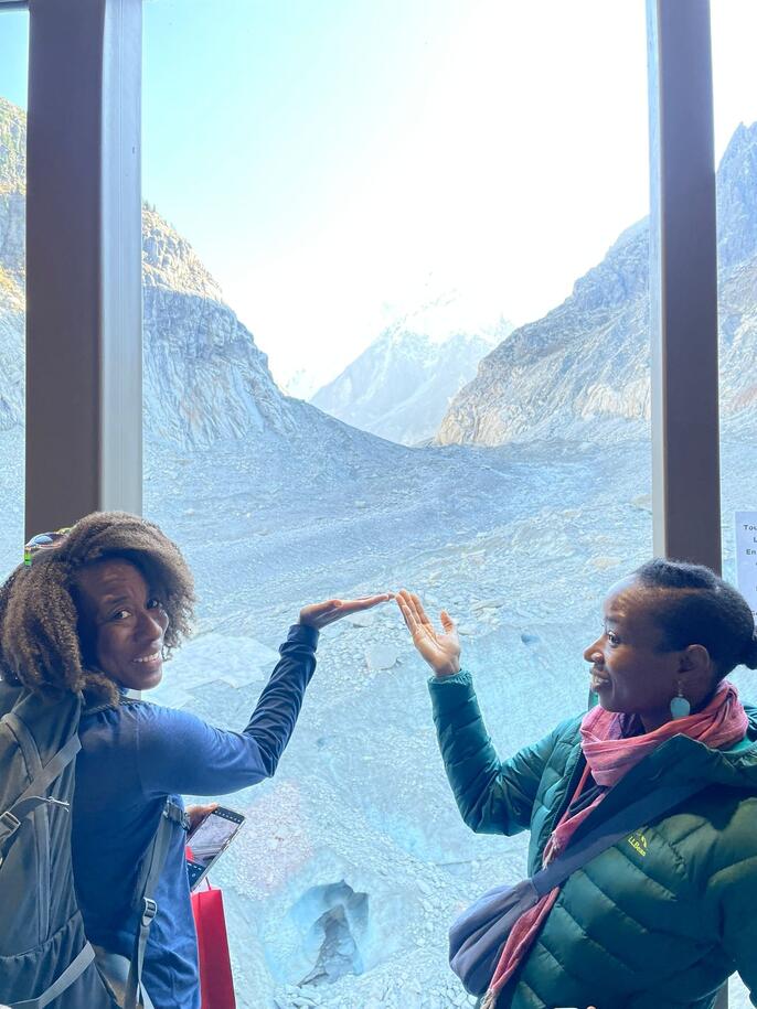 Jemise Ray De La Cruz and her sister smile and gesture toward the alpine landscape visible through a large window overlooking the Mont Blanc mountain range.