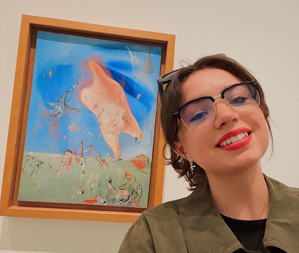 A young woman wearing two pairs of glasses smiling in front of Salvador Dali's painting titled "Sterile Efforts"
