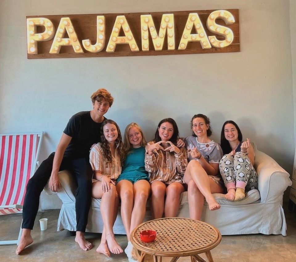 6 people in pajamas smile and pose on a white couch beneath a lit-up sign that says "Pajamas"