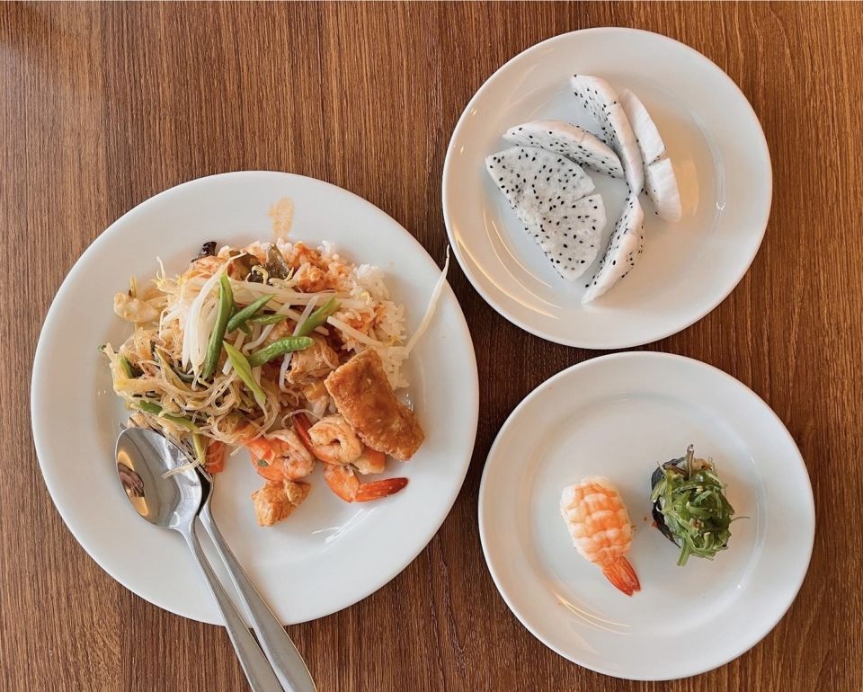 Three white plates filled with sushi, pad thai, and dragon fruit on a brown wooden table