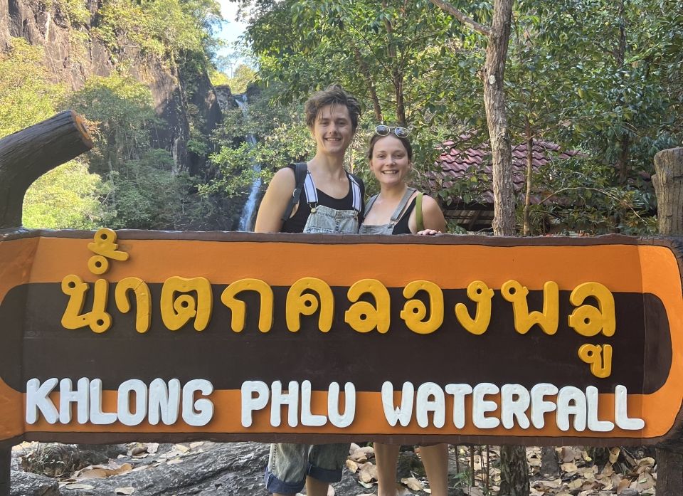 Me and my boyfriend smiling in front of a waterfall with the sign "Khlong Phlu Waterfall"