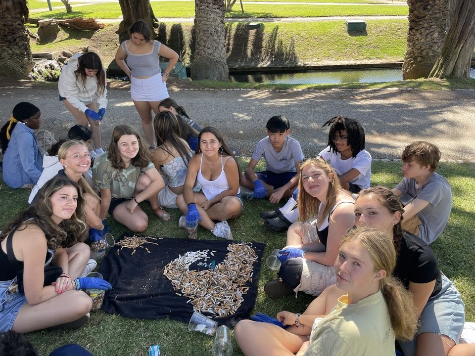 Students sit on the grass around a black towel with a wave made of cigarettes on it.