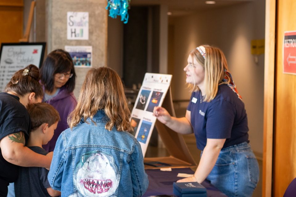 A teenaged woman across a table explaining something to a group of younger students