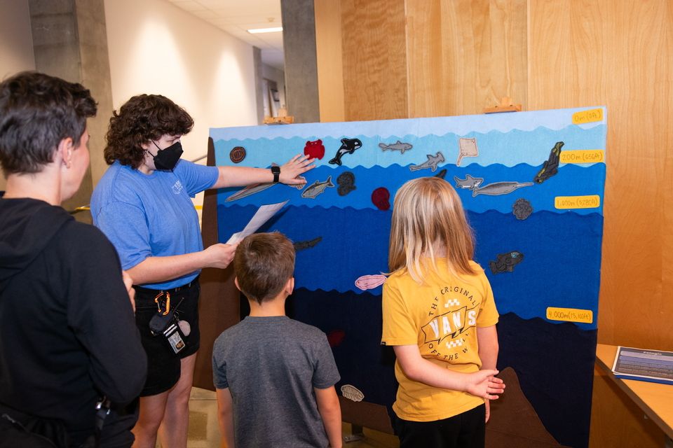 3 young students looking at a display image of the ocean with an older teenage student pointing to the display while explaining a topic