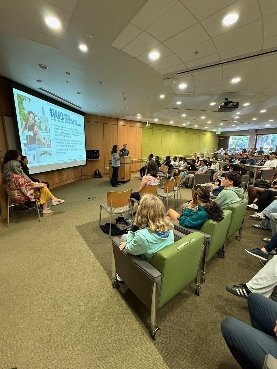 a wide shot of a classroom with students and parents looking at a projector screen with a slide on it