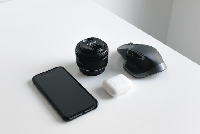 Photo of technology: a phone, airpods, a wireless mouse, and a DSLR camera lens