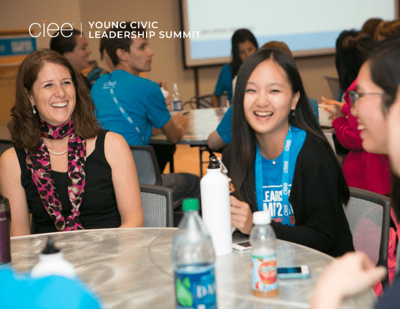 A young student sits mid-laugh at a table with a baby blue shirt on that says "Young Civic Leadership Summit"